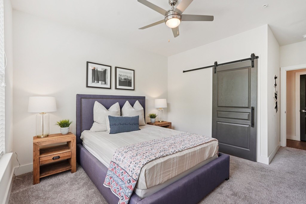 A  large bedroom with a bed, nightstands, ceiling fan., and a walk-in closet with a barn door